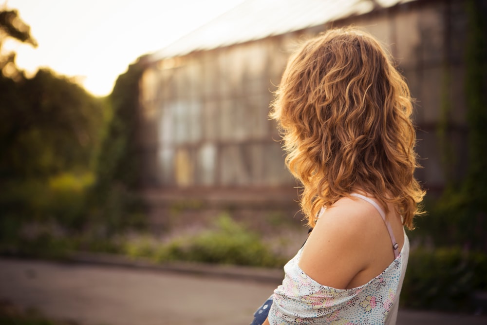 A woman with wavy hair and off-the-shoulder top looks away toward a greenhouse